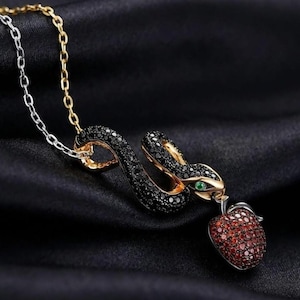Puede incluir: Un collar con un colgante de serpiente en tono dorado incrustado con piedras negras y un ojo verde, que sostiene un dije de manzana pavimentado con piedras rojas. El colgante cuelga de una cadena bicolor con eslabones plateados y dorados.