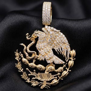 May include: A gold-colored pendant featuring the Mexican coat of arms, including an eagle with a snake in its beak, surrounded by a wreath. The pendant is encrusted with small, sparkling stones and has a diamond-studded bail.