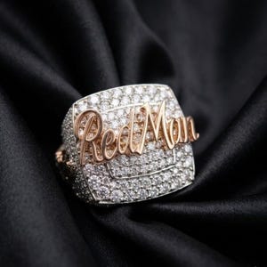May include: A square-faced ring with a silver band and a gold-colored script that reads "Red Man". The ring is encrusted with numerous small, clear stones. The ring is set against a black background.