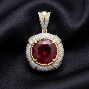 May include: A circular pendant featuring a large, round, deep red gemstone at the center, surrounded by a gold rope-style border and an outer frame encrusted with numerous small, clear white stones, finished with a diamond-shaped bail.