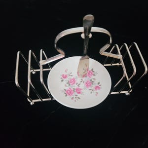 Puede incluir: Un soporte para tostadas de metal plateado con un plato central circular de cerámica blanca decorado con un patrón de rosas rosas y hojas verdes. Un cuchillo de untar de metal plateado está colocado verticalmente en el centro.