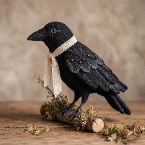 May include: A needle-felted black crow figurine perched on a small wooden branch covered in green moss. The crow features a textured black body, a dark beak, a single visible eye, and a decorative cream-coloured lace ribbon tied around its neck.