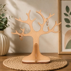 Pode incluir: Um suporte para joias de madeira de cor clara em forma de árvore com múltiplos ramos para pendurar objetos. O suporte apresenta uma base plana e estável e um tronco central, com dois anéis metálicos finos pendurados nos ramos.