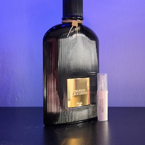 May include: A black Tom Ford Black Orchid perfume bottle with a ribbed design and gold accents. A smaller sample bottle is next to it. The large bottle has a gold label that reads "Tom Ford Black Orchid 100 ML".