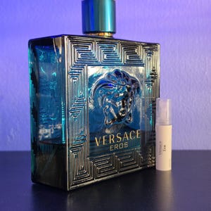 May include: A turquoise Versace Eros fragrance bottle with a silver geometric design and a matching sample vial. The bottle has a gold and turquoise cap and the Versace logo is visible on the front.