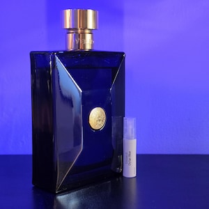 May include: A dark blue rectangular perfume bottle with a gold cap and a gold emblem. A small white vial with the text "Versace Dylan Blue" is next to the bottle. The background is a gradient of blue.