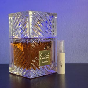 May include: A square glass perfume bottle with a textured, faceted surface containing amber liquid, featuring a gold label with the text Khamrah Lattafa. Beside it stands a small, clear plastic sample vial labelled Lattafa Khamrah.