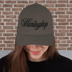 Op de afbeelding: Een donkergrijze baseballpet met het woord Haringkop in zwarte schrijfletters op de voorzijde geborduurd. De pet heeft een gestructureerde kroon, een gebogen klep en een klein zwart stersymbool dat boven de letter i is geplaatst.
