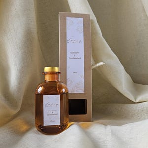 May include: An amber-colored diffuser bottle with a gold cap, labeled "Mandarin & Sandalwood." A matching box stands behind it, also labeled with the scent and brand name. The background is a neutral-toned fabric.