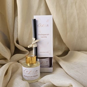 Peut inclure: Un flacon en verre transparent avec un bouchon doré et une étiquette blanche portant l'inscription "Sofia Dourvari Madagascan Vanilla Reed Diffuser". À côté, une boîte blanche avec le même texte et un bouquet de bâtonnets noirs.