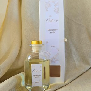 May include: A clear glass diffuser bottle with a gold cap and a white label that reads "Madagascan Vanilla". A white box with the same text and floral design is behind the bottle. The background is a beige fabric.