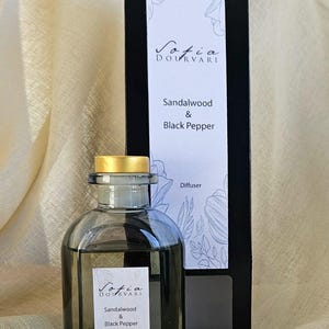 May include: A clear glass diffuser bottle with a gold cap, filled with a dark liquid. A black box with the text "Sofia Dourvari" and "Sandalwood & Black Pepper" is behind the bottle. The product is a diffuser.