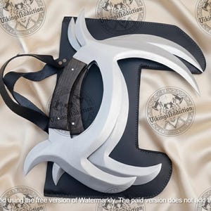 May include: A set of silver-colored, curved blades with a dark wooden handle, resting on a black leather sheath. The blades are arranged in a symmetrical design. A black strap is attached to the sheath. The text "Vikings Addiction" is visible.