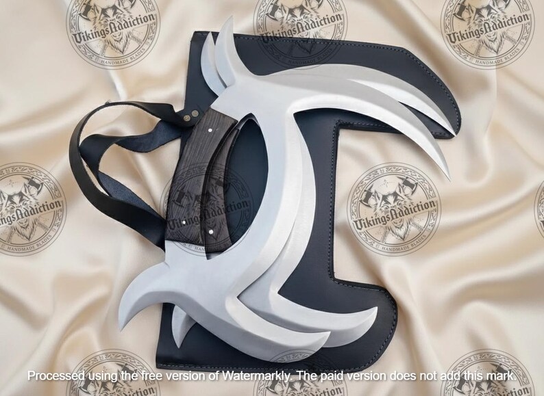 May include: A set of silver-colored, curved blades with a dark wooden handle, resting on a black leather sheath. The blades are arranged in a symmetrical design. A black strap is attached to the sheath. The text "Vikings Addiction" is visible.