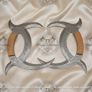 May include: Two curved, silver-colored blades with a patterned design and wooden handles. The blades are arranged symmetrically on a cream-colored fabric. The text "VikingsAddiction" is visible.