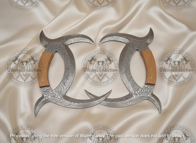 May include: Two curved, silver-colored blades with a patterned design and wooden handles. The blades are arranged symmetrically on a cream-colored fabric. The text "VikingsAddiction" is visible.