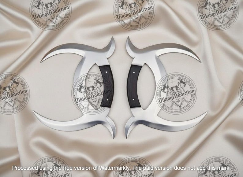 May include: Two silver-colored, curved blades with black handles. The blades have a crescent shape and are displayed on a cream-colored, satin-like surface. The handles are dark with a textured appearance.