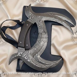 May include: A Damascus steel karambit knife with a dark wooden handle and a black leather sheath. The blade has a unique curved design. The knife is displayed on a cream-colored fabric.