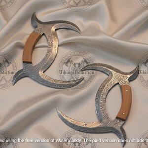 May include: Two curved, Damascus steel blades with wooden handles. The blades have a unique pattern and are displayed on a satin surface. The handles are light brown. The text "Vikings Addiction" is visible.