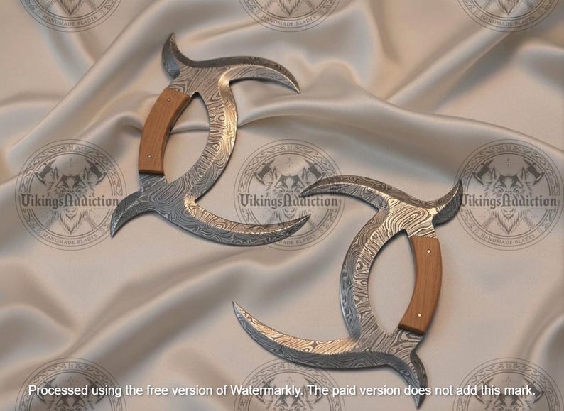 May include: Two curved, Damascus steel blades with wooden handles. The blades have a unique pattern and are displayed on a satin surface. The handles are light brown. The text "Vikings Addiction" is visible.