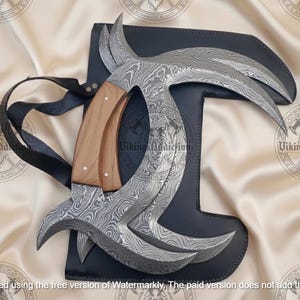 May include: A Damascus steel karambit knife with a wooden handle rests on a black leather sheath. The blade has a curved design with a unique pattern. A black strap is attached to the sheath. The knife is a handmade item.