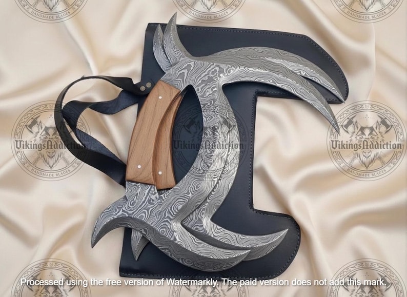 May include: A Damascus steel karambit knife with a wooden handle rests on a black leather sheath. The blade has a curved design with a unique pattern. A black strap is attached to the sheath. The knife is a handmade item.
