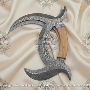 May include: A Damascus steel karambit knife with a curved blade and a wooden handle. The blade has a unique pattern, and the handle is light brown. The knife is displayed on a cream-colored fabric.