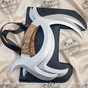 May include: A handmade Viking-style blade with a curved, silver-colored blade and a wooden handle. The blade is set in a black leather sheath with a black strap. The background is a cream-colored fabric with repeating logos.