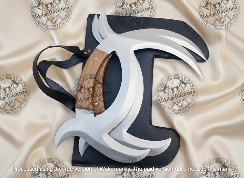 May include: A handmade Viking-style blade with a curved, silver-colored blade and a wooden handle. The blade is set in a black leather sheath with a black strap. The background is a cream-colored fabric with repeating logos.