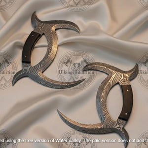 May include: Two Damascus steel karambit knives with curved blades and dark wood handles. The blades have a unique pattern, and the handles are riveted. The knives are displayed on a satin fabric.