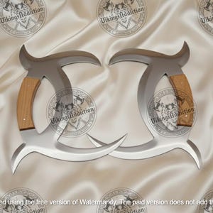 May include: Two silver-colored, curved blades with wooden handles. The blades have a unique shape with a pointed tip and a curved top. The handles are made of light brown wood. The background is a white, silky fabric with circular logos.
