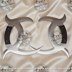 May include: Two silver-colored, curved blades with black wooden handles. The blades have a unique shape with a hook at the top and a pointed end. The blades are displayed on a white, textured surface.