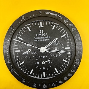 May include: Black Omega Speedmaster Professional watch face with white hands and markings. Features sub-dials, a tachymeter scale, and the Omega logo. The watch face is set against a yellow background.