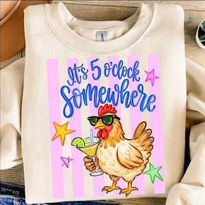 May include: A cream-coloured sweatshirt featuring the text "It's 5 o'clock Somewhere" in blue script. The design includes a cartoon chicken wearing sunglasses and holding a cocktail, with pink stripes and star accents.