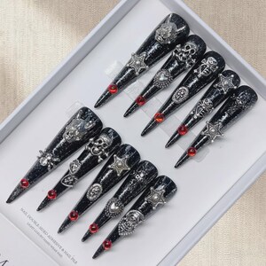 May include: A set of black glittered, long, pointed false nails with silver gothic-style embellishments. Each nail features a different design, including skulls, stars, and ornate filigree, with a small red gem near the tip. The nails are displayed in a white box.