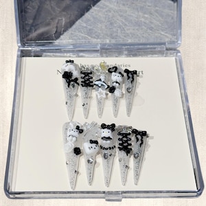 May include: A set of ten long, clear acrylic nails with various black and white decorative elements. Each nail features a different design, including bows, cartoon characters, and sparkling accents. The nails are displayed in a clear plastic case.
