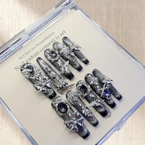 Puede incluir: Un conjunto de diez uñas postizas largas en forma de ataúd. Cada uña es de color gris oscuro con un efecto ojo de gato, adornada con adornos plateados que incluyen gemas, perlas y piezas metálicas decorativas. El texto "Art has no boundaries Nail enhance cement is art" es visible.