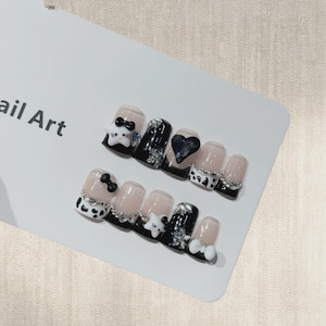 May include: A set of artificial nails with various designs. Some nails are black, others are nude, and some feature a Hello Kitty design. Other nails have a heart, star, and bow accents. The nails are decorated with rhinestones and other embellishments.