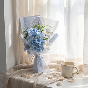 Op de afbeelding: Een boeket handgemaakte blauwe en witte bloemen, verpakt in lichtblauw papier en vastgebonden met een lint. Het boeket staat op een beige oppervlak naast een witte mok en schelpen. De bloemen omvatten hortensia's en madeliefjes.