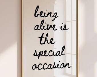 May include: A rectangular wall art print in a thin gold frame featuring the black handwritten text being alive is the special occasion on a light beige background. The artwork is displayed in a vertical orientation with clean lines.