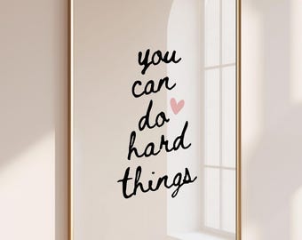 May include: A rectangular wall art print in a thin gold frame featuring the black handwritten text you can do hard things with a small light pink heart icon positioned between the words do and hard on a plain off-white background.