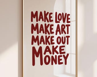 May include: A rectangular wall art print in a thin gold frame featuring dark red text on a light beige background. The text reads: MAKE LOVE, MAKE ART, MAKE OUT, MAKE MONEY. The lettering has a hand-painted, brushstroke style with uneven edges.