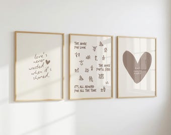 May include: Three framed wall art prints with light wood frames. The left print says 'love's never wasted when it's shared.' The middle print features small line drawings of people and the text 'THE MORE YOU LOOK THE MORE YOU'LL FIND IT'S ALL AROUND YOU ALL THE TIME.' The right print shows a large brown heart.