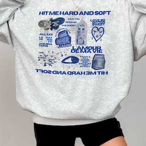 May include: A light grey sweatshirt with blue text and graphics. The design includes the words "HIT ME HARD AND SOFT", "L'AMOUR DE MA VIE", and "DINER". The sweatshirt is a casual style.