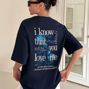 May include: Navy blue t-shirt with the text "i know that you love me" in white and blue. The shirt features floral accents and additional text. The shirt is a casual wear item.