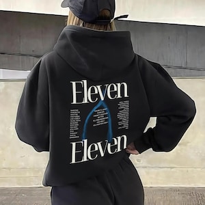 Op de afbeelding: Zwarte hoodie met het woord "Eleven" in het wit, met extra tekst en een blauwe afbeelding op de achterkant. De hoodie heeft een capuchon met trekkoord en een relaxte pasvorm. De persoon draagt een zwarte pet.
