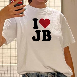 Puede incluir: Camiseta blanca con el texto "I ❤️ JB" en negro y un corazón rojo. La camiseta informal probablemente está hecha de algodón o un material similar.
