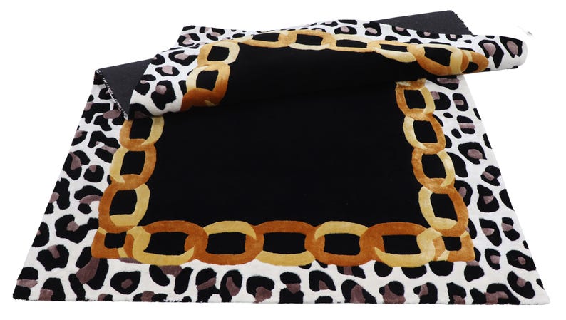 May include: A rectangular area rug featuring a central solid black square bordered by a gold-toned chain link pattern. The outer edge displays a white background with a black and brown leopard print motif, with one corner folded over to show the back.