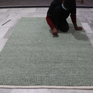Puede incluir: Una alfombra rectangular verde salvia con una superficie texturizada y un borde color crema. La alfombra está hecha de un material tejido y se exhibe sobre un suelo de mármol. La alfombra mide aproximadamente 152 cm de largo.