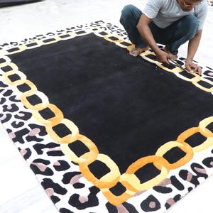 May include: A rectangular area rug featuring a solid black centre, a border composed of interlocking golden yellow chain links, and an outer edge patterned with black and light brown leopard spots on a white background.